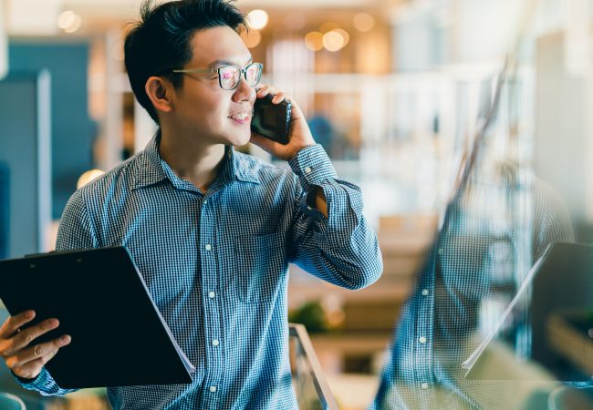 A young asian man holding a tablet and speaking on the phone at the office