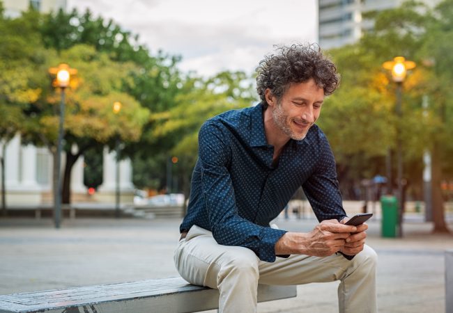 Mature businessman sitting on bench and using smart phone at dusk. Happy smiling guy in smart casual reading a message on cellphone. Stylish man surfing the net with smart phone in the evening.