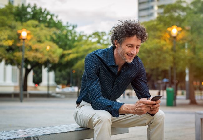 Mature businessman sitting on bench and using smart phone at dusk. Happy smiling guy in smart casual reading a message on cellphone. Stylish man surfing the net with smart phone in the evening.