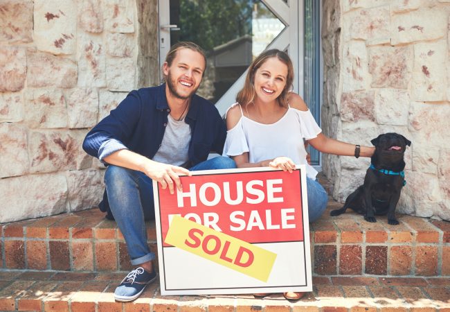 Portrait of a young couple holding a for sale sign while sitting outside their new home