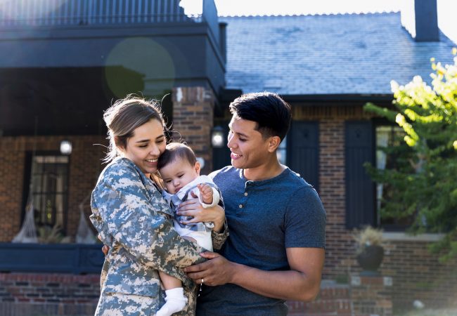 Military wife holding young son and standing next to husband loving standing in front of their home