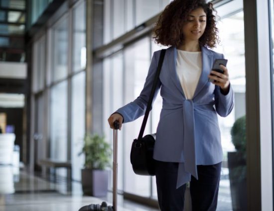 Business woman in blue blazer walking through a building holding a cellphone and rolling a suitcase behind her