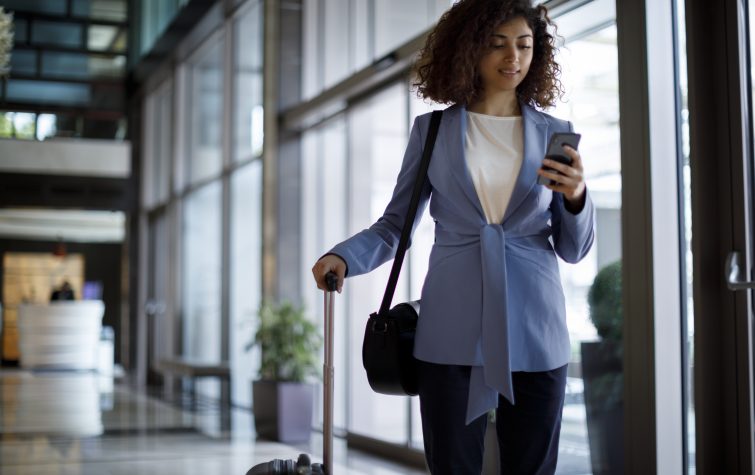 Business woman in blue blazer walking through a building holding a cellphone and rolling a suitcase behind her