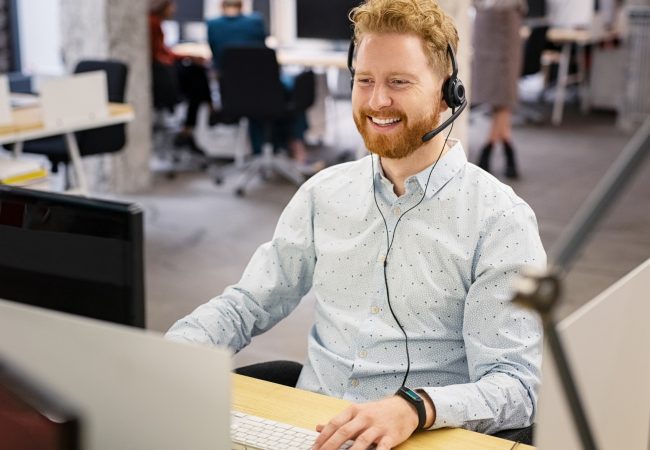 Portrait of consultant agent man in call center smiling. Happy customer support agent working with headset while sitting at his workstation. Smiling telephone operator using computer in modern open space office.