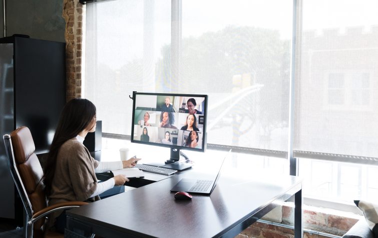 A group of creative professionals meet virtually to discuss and brainstorm ideas. A young female creative professional is working in an office while talking with colleagues who are telecommuting from their homes.