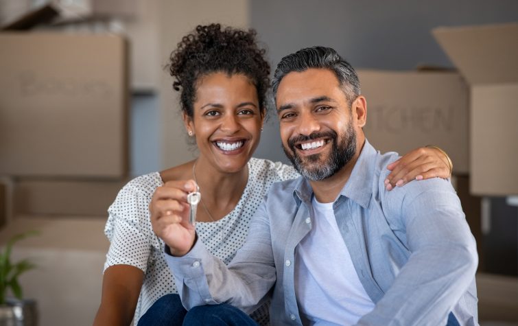 Couple sitting on floor holding house keys with carboard boxes behind them.