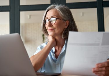 Woman similing in front of a laptop, holding a piece of paper and looking out of a window