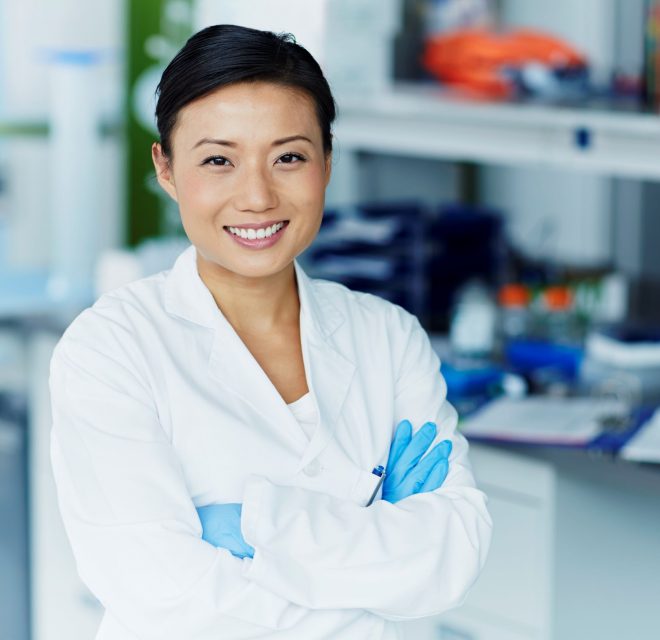 Woman dressed in lab coat and gloves smiling with her arms crossed