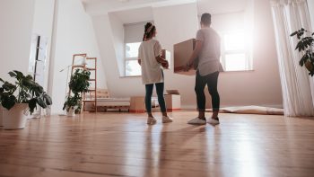 Full length rear view shot of young couple carrying cardboard box at new home. Young man and woman holding boxes and moving in new house.