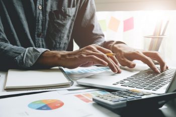 Close up of businessman or accountant hand holding pen working on laptop computer for calculate business data, accountancy document and calculator at office