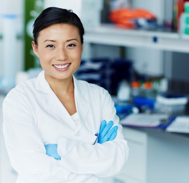 Woman dressed in lab coat and gloves smiling with her arms crossed