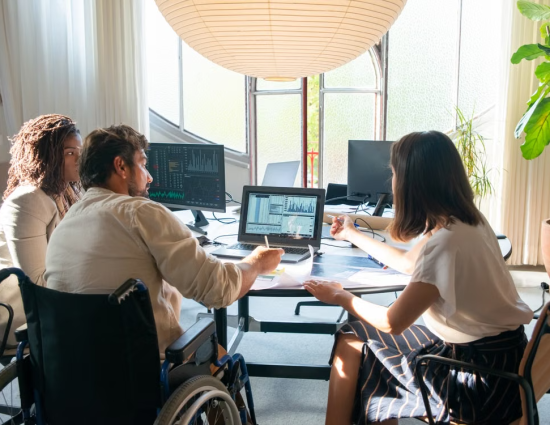 Diverse team of three professionals including a wheelchair user reviewing mobility program data and analytics on multiple screens in a modern office
