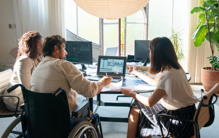 Diverse team of three professionals including a wheelchair user reviewing mobility program data and analytics on multiple screens in a modern office
