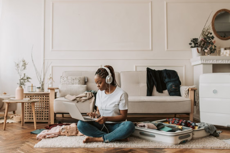 Woman wearing headphones using a laptop beside an open suitcase in a living room representing a relocating employee planning a temporary housing stay